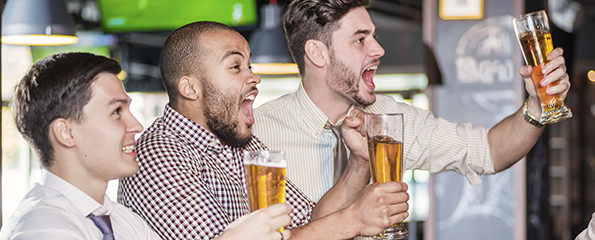 Men fans screaming and watching football on TV and drink beer. Three other men drinking beer and having fun together in the bar until the bartender standing near the rack